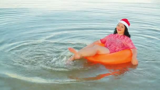 Brunetka w kręgu pływackim i kapelusz Świętego Mikołaja spędzają Boże Narodzenie i Nowy Rok na morzu. — Wideo stockowe