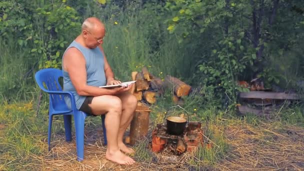 Een man bij het vuur leest een boek en kookt eten in een pot.. — Stockvideo