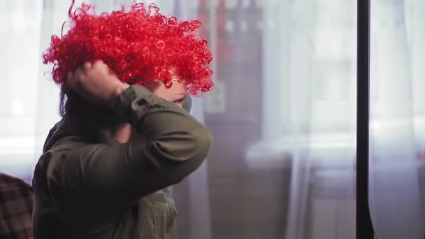Een vrouw voor een spiegel zet een clown pruik op met rode krullen. — Stockvideo