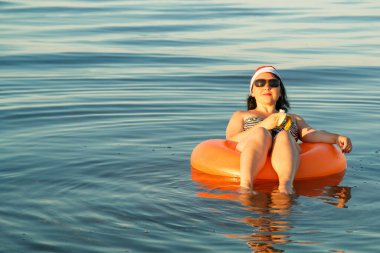 Esmer bir kadın yüzme havuzunda yatıyor ve Noel Baba şapkasıyla muz yiyor..