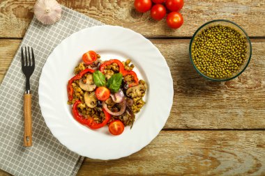 Ahşap bir masada, içinde mantar ve sebzeler olan kaynamış bir fasulye tabağı ve malzemelerin yanında da bir çatal var..