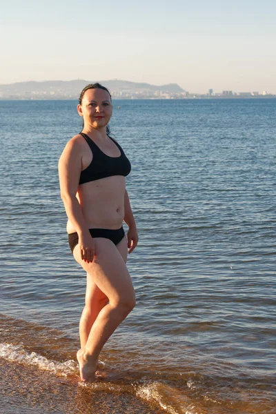 Une femme en maillot de bain noir marche le long du littoral le matin. — Photo