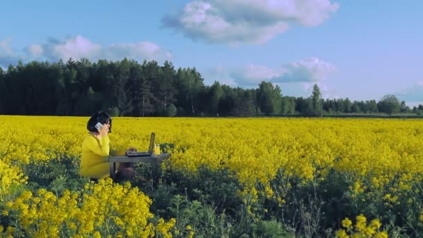 Μια γυναίκα με κίτρινα ρούχα στη μέση ενός αγρού με κίτρινα λουλούδια δουλεύει εξ αποστάσεως σε έναν υπολογιστή και πίνει χυμό και μιλάει στο τηλέφωνο.. — Αρχείο Βίντεο