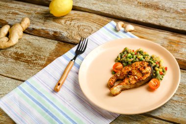 Keten peçetenin üzerinde pişmiş alabalık bifteği, yeşil sebzeli kinoa ve yanında çatal olan bir tabak var..