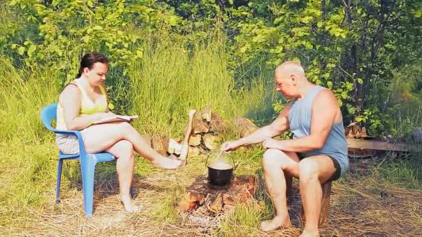 Een man bij het vuur kookt eten in een pot in een pot, en zijn vrouw leest een boek — Stockvideo