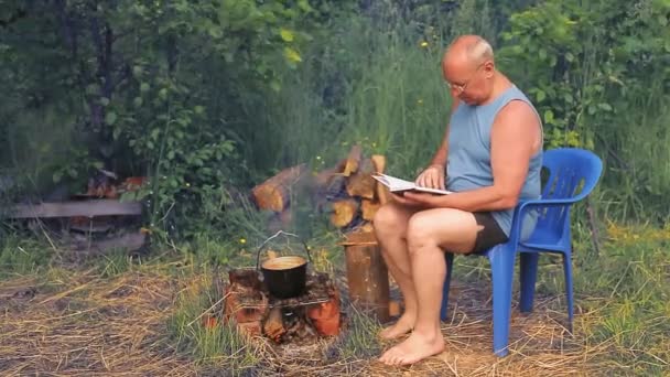 Een man bij het vuur leest een boek en kookt eten in een pot.. — Stockvideo