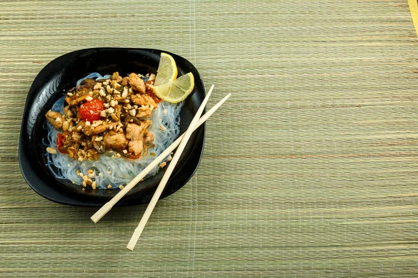 Prato com funchose e frango com legumes em pad molho tailandês ao lado de pauzinhos em uma esteira de bambu . — Fotografia de Stock