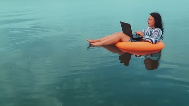 Μια νεαρή γυναίκα με ένα λαστιχένιο δαχτυλίδι κολυμπάει κοντά στη θάλασσα το πρωί και δουλεύει σε ένα λάπτοπ.. — Αρχείο Βίντεο
