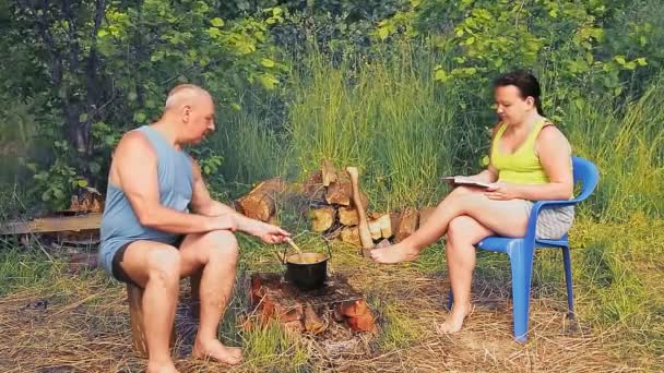 Een man bij het vuur kookt eten in een pot in een pot, en zijn vrouw leest een boek — Stockvideo