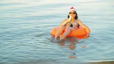 Mayo giymiş esmer bir kadın, yüzme çemberinde güneş gözlüğü ve Noel Baba şapkası takan bir kadın Noel 'de ve yılbaşında denizde muz yiyor..