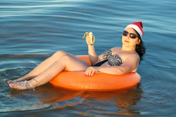 Брюнетка в купальнике и шляпа Санта Клауса в круге купания плавает в море. — стоковое фото