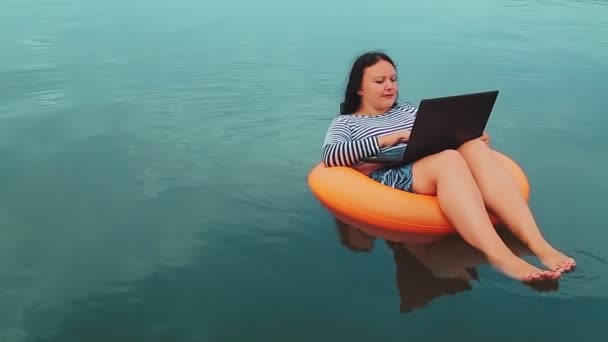 Uma jovem freelancer em um anel inflável nada perto do mar pela manhã e trabalha em um laptop — Vídeo de Stock