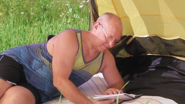 Mężczyzna w otwartym namiocie kłamie i czyta książkę w słoneczny dzień na polanie.. — Wideo stockowe