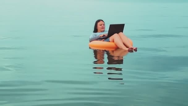 Junge Geschäftsfrau im Urlaub schwimmt morgens im Gummiring am Meer und arbeitet in einem Laptop — Stockvideo