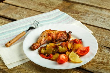 Ahşap bir masada çatalla birlikte beyaz bir tabakta patates ve domatesli fırında tavuk budu..