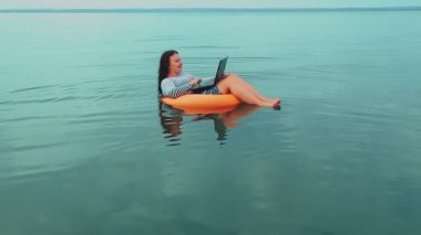 Lastik halkalı genç bir kadın sabah denize yakın yüzer ve dizüstü bilgisayarda çalışır..