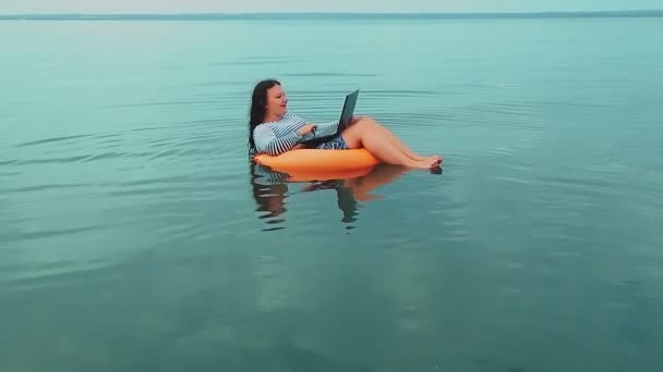En ung kvinna i en gummiring simmar nära havet på morgonen och arbetar i en bärbar dator. — Stockvideo