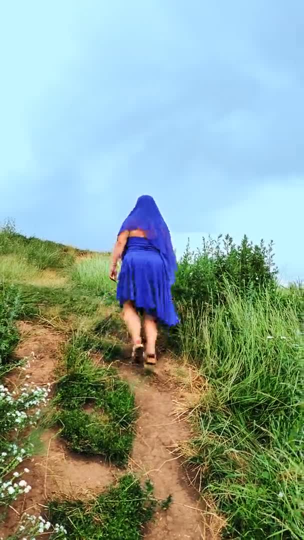 Una mujer peregrina en una capa sobre su cabeza en azul sube a la montaña hasta la cruz. Vertical — Vídeo de stock