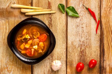 Geleneksel Tayland çorbası Tom Yam, ahşap bir masada, sarımsak ve limon otunun yanında, kaffir limonu, kırmızı biber ve kiraz..
