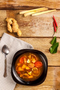 Geleneksel Tayland Tom Yam çorbası, siyah bir tabakta, tahta bir masada, yanında kaşıkla peçetenin üstünde..