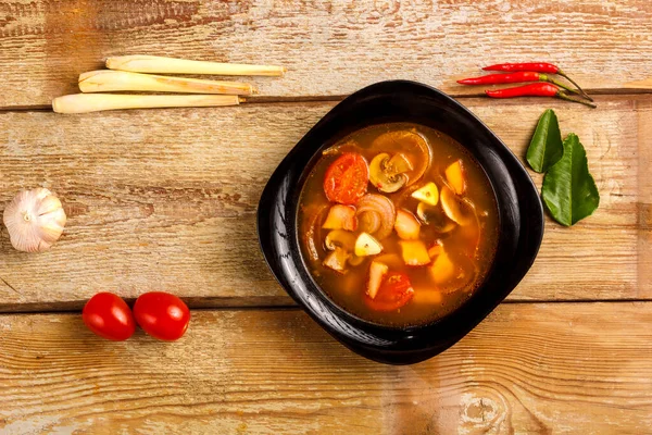 Traditionele Thaise soep Tom Yam in een bord op een houten tafel naast knoflook en citroengras, kaffirlimoen, chili en kers. — Stockfoto