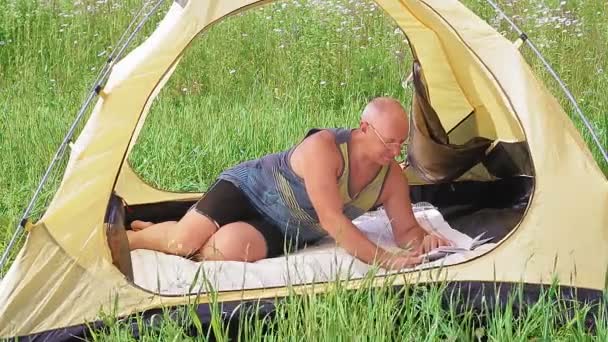 En man i ett öppet tält ligger och läser en bok en solig dag i en glänta. — Stockvideo