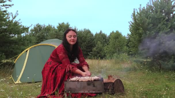 Een jonge slimme zigeunerin op een open plek bij de tent grilt vlees op de grill. — Stockvideo