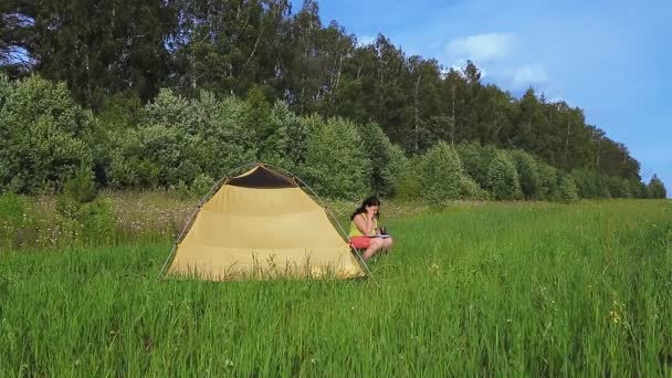 Uma mulher perto de uma tenda em uma clareira lê um livro e bebe chá . — Vídeo de Stock