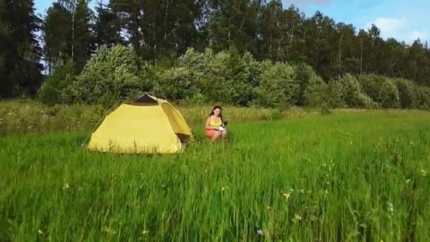 Kobieta w pobliżu namiotu w zielonej polanie pije herbatę — Wideo stockowe