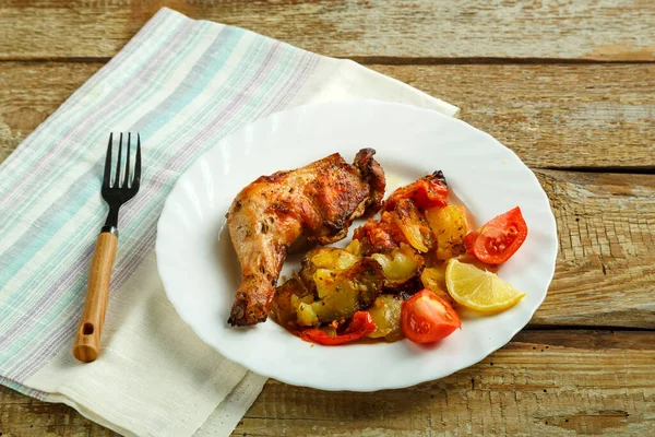 烤鸡腿，白盘上有土豆和西红柿，木桌上有叉子. — 图库照片
