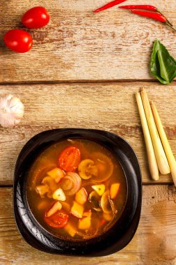 Geleneksel Tayland çorbası Tom Yam, ahşap bir masada, sarımsak ve limon otunun yanında, kaffir limonu, kırmızı biber ve kiraz..