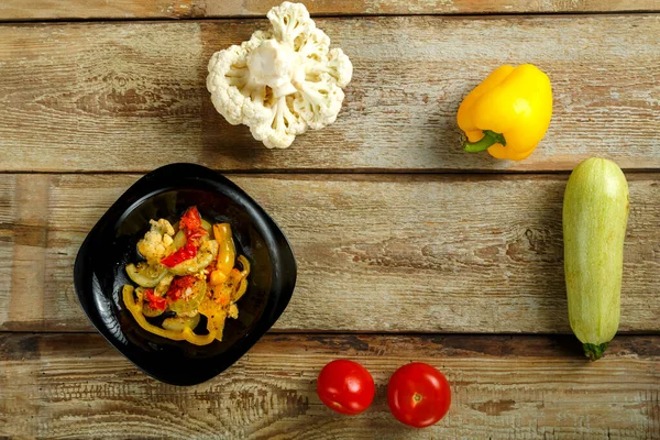 Ragoût de légumes sur une assiette noire et plusieurs légumes sur une table en bois. — Photo