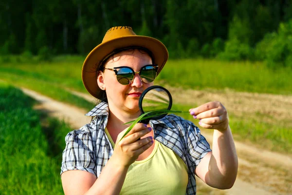 Uma mulher agrônomo em óculos de sol e um chapéu examina os brotos de culturas em uma lupa . — Fotografia de Stock