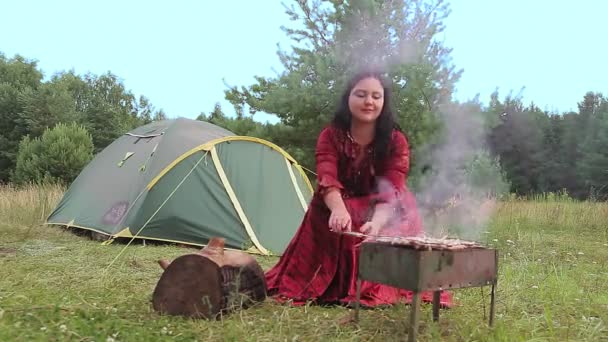 Молодая умная цыганка жарит мясо на поляне у палатки.. — стоковое видео