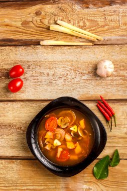 Geleneksel Tayland çorbası Tom Yam, ahşap bir masada, sarımsak ve limon otunun yanında, kaffir limonu, kırmızı biber ve kiraz..