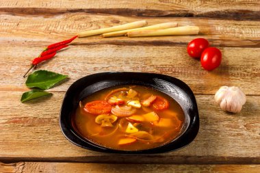 Geleneksel Tayland çorbası Tom Yam, ahşap bir masada, sarımsak ve limon otunun yanında, kaffir limonu, kırmızı biber ve kiraz..