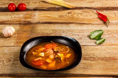 Geleneksel Tayland çorbası Tom Yam ahşap bir masada, sarımsak ve limon otunun yanında, kaffir limonu, kırmızı biber ve kiraz..