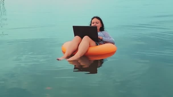 Una joven freelancer en un anillo inflable nada cerca del mar por la mañana y trabaja en un portátil . — Vídeo de stock