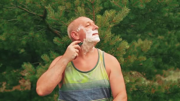 Ένας άντρας στο δάσος σε ένα ξέφωτο ξυρίζει το πρόσωπό του με αφρό με ξυράφι.. — Αρχείο Βίντεο