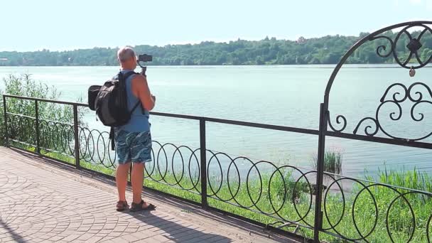 Un blogueur masculin avec un téléphone portable parle de voyage sur le remblai d'une petite ville. — Video