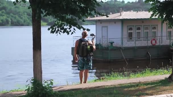 Un blogger masculino camina por la orilla del río con un teléfono móvil y bloguea — Vídeo de stock