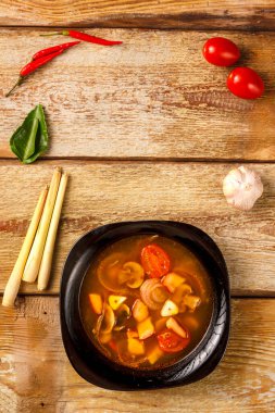 Geleneksel Tayland çorbası Tom Yam, ahşap bir masada, sarımsak ve limon otunun yanında, kaffir limonu, kırmızı biber ve kiraz..