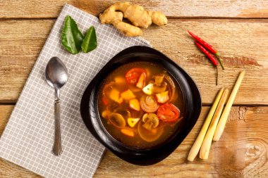 Geleneksel Tayland Tom Yam çorbası, siyah bir tabakta, tahta bir masada, yanında kaşıkla peçetenin üstünde..