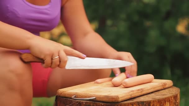 Mujer corta salchichas con un cuchillo en una tabla de madera en un picnic. — Vídeos de Stock