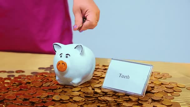 Uma mão de mulher põe uma moeda de porco em um banco porquinho próximo a moedas e os dentes de inscrição . — Vídeo de Stock