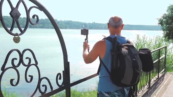 Un blogger masculino con un teléfono móvil sobre viajes habla sobre el terraplén de un gran río. — Vídeos de Stock
