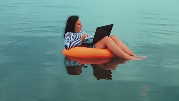 Μια νεαρή γυναίκα ελεύθερη επαγγελματίας σε ένα φουσκωτό δαχτυλίδι κολυμπά κοντά στη θάλασσα το πρωί και εργάζεται σε ένα φορητό υπολογιστή — Αρχείο Βίντεο