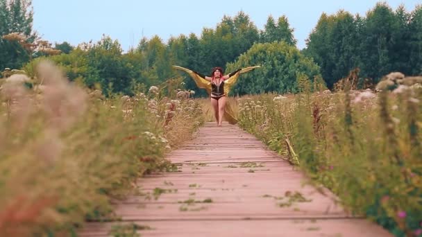En kvinna i form av en demon i svart korsett med gyllene vingar löper längs en träväg — Stockvideo