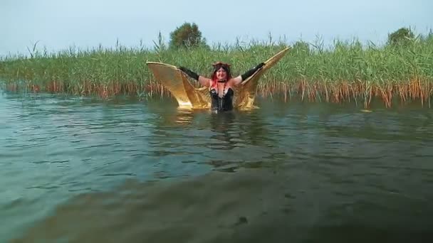 Een vrouw in de vorm van een demon in een zwart korset met gouden vleugels komt uit het water van het meer en klapt met haar vleugels. — Stockvideo