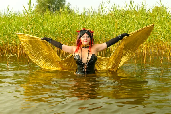 A woman in the image of a demon in a black corset with golden wings in the water in the lake with fluttering wings in the reeds. — Stock Photo, Image